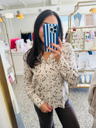A woman takes a mirror selfie in a boutique wearing a beige knit top with a black leopard print pattern and dark brown faux-leather pants. She is holding a blue and white striped phone.