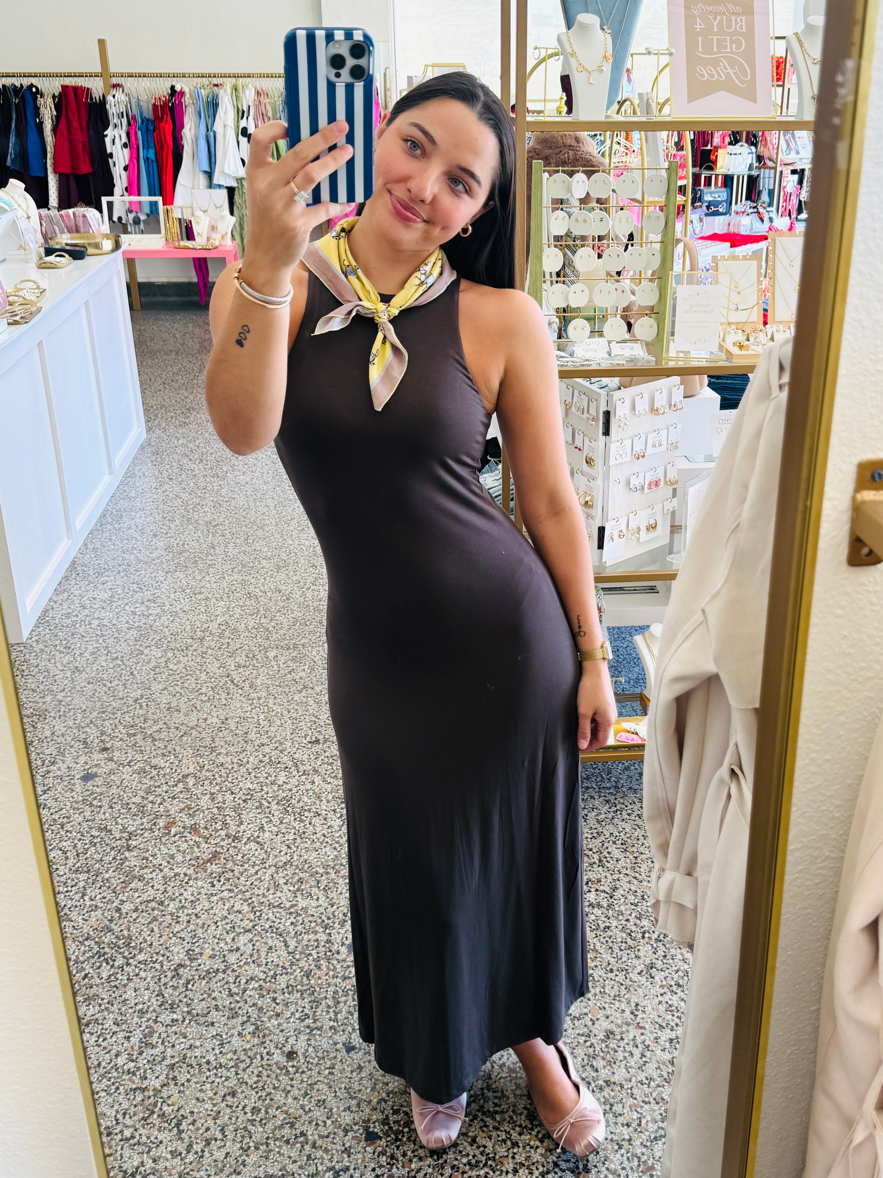 A woman takes a mirror selfie in a boutique wearing a sleeveless, dark brown halter-neck maxi dress with a patterned yellow and white neck scarf. The boutique interior features a speckled floor, clothing racks, and display shelves.