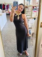 A woman takes a mirror selfie in a boutique wearing a sleeveless, dark brown halter-neck maxi dress with a patterned yellow and white neck scarf. The boutique interior features a speckled floor, clothing racks, and display shelves.