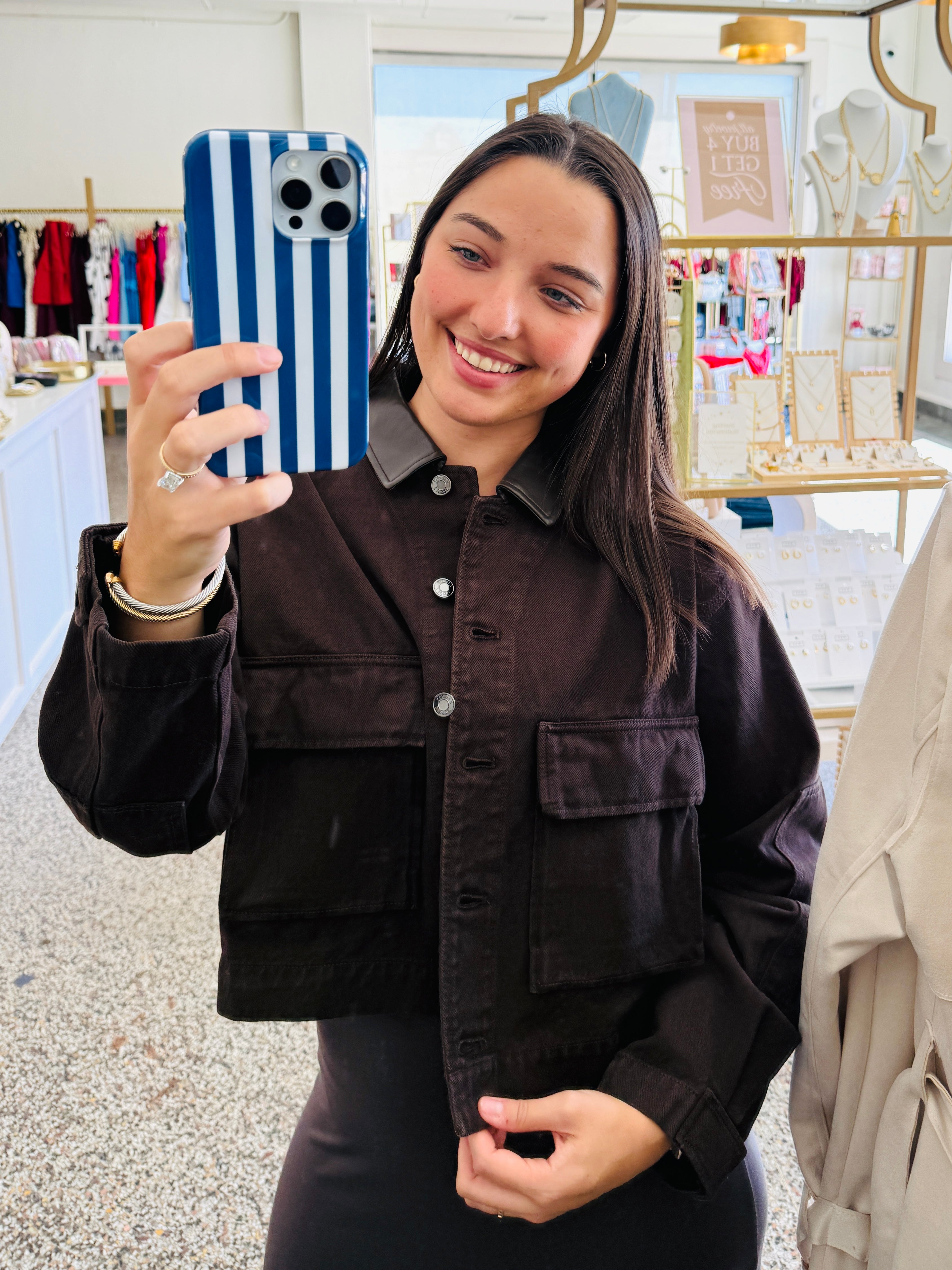A woman taking a mirror selfie in a boutique, wearing a dark brown corduroy button-up shirt jacket with a pointed collar and front flap pockets.