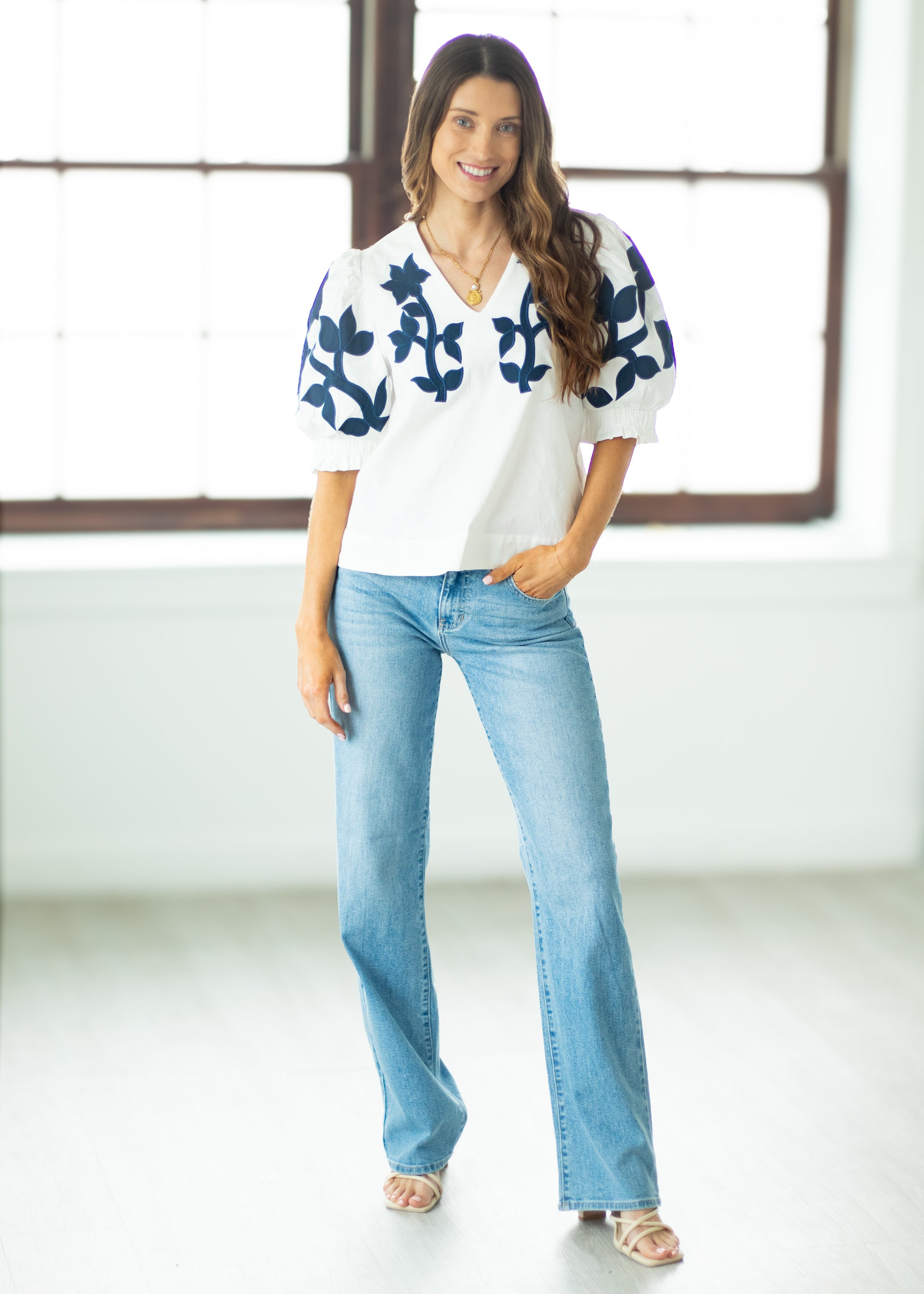 Woman wearing a white blouse with blue patterns and blue jeans standing in a bright room.