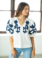 Woman wearing a white blouse with blue floral patterns and blue jeans indoors.
