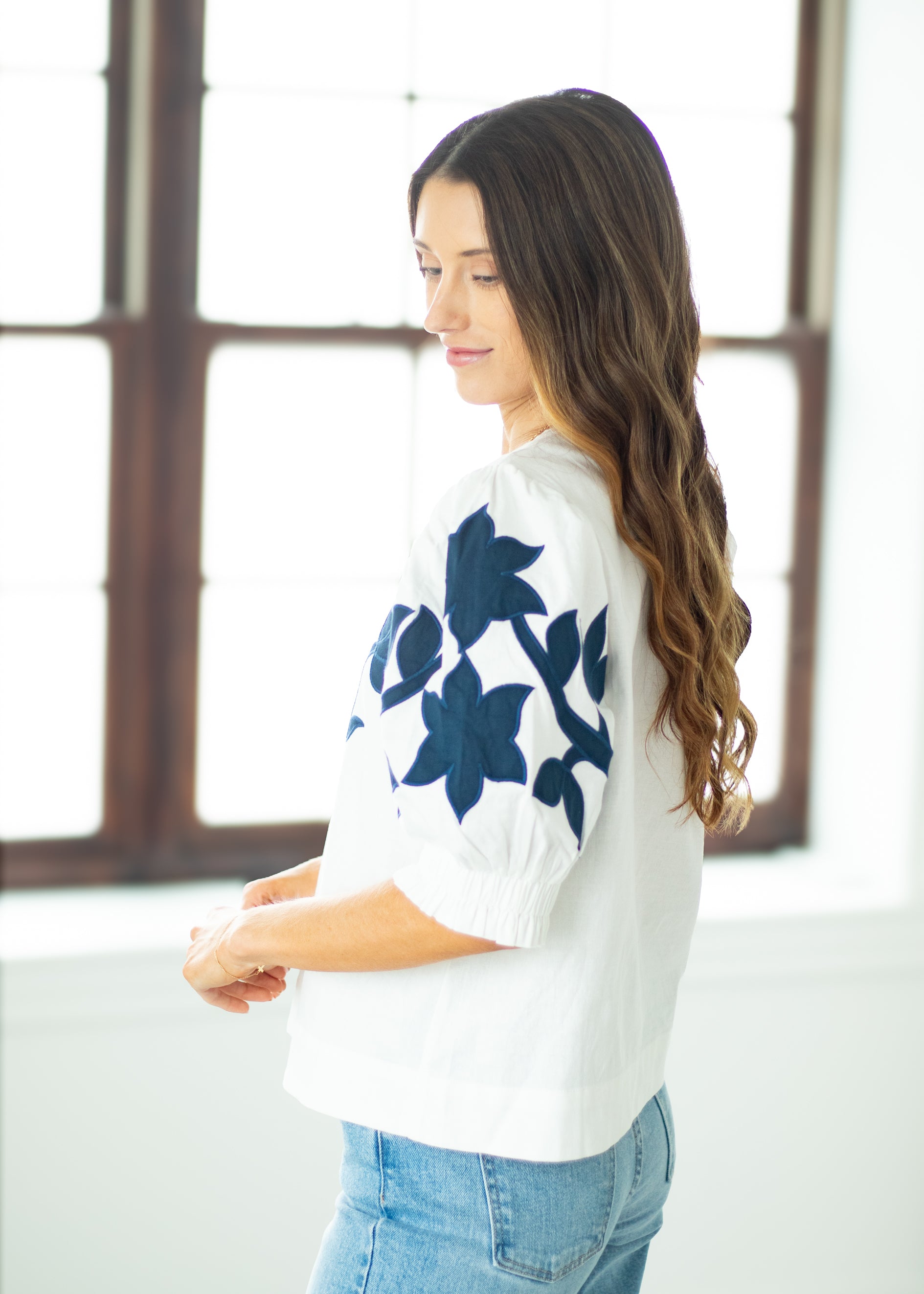 Woman wearing a white sweater with blue floral patterns in a bright room.
