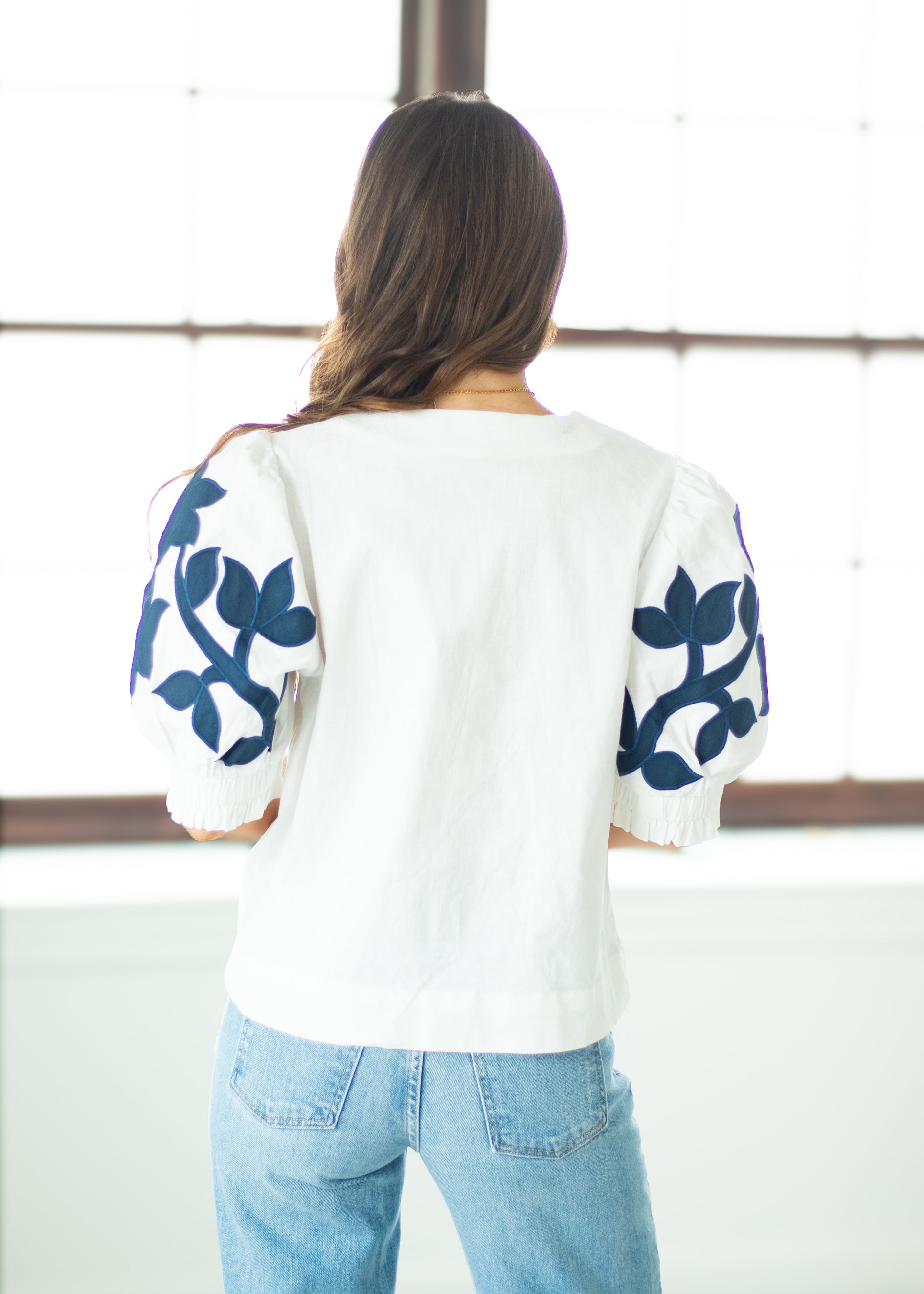 Woman wearing a white top with blue floral patterns and blue jeans, standing against a white wall.