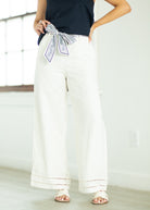 Person wearing white wide-leg pants with a navy top and decorative ribbon, standing against a light background.