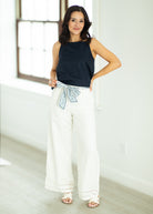 Woman wearing a black sleeveless top and white pants with a decorative belt in a bright room.