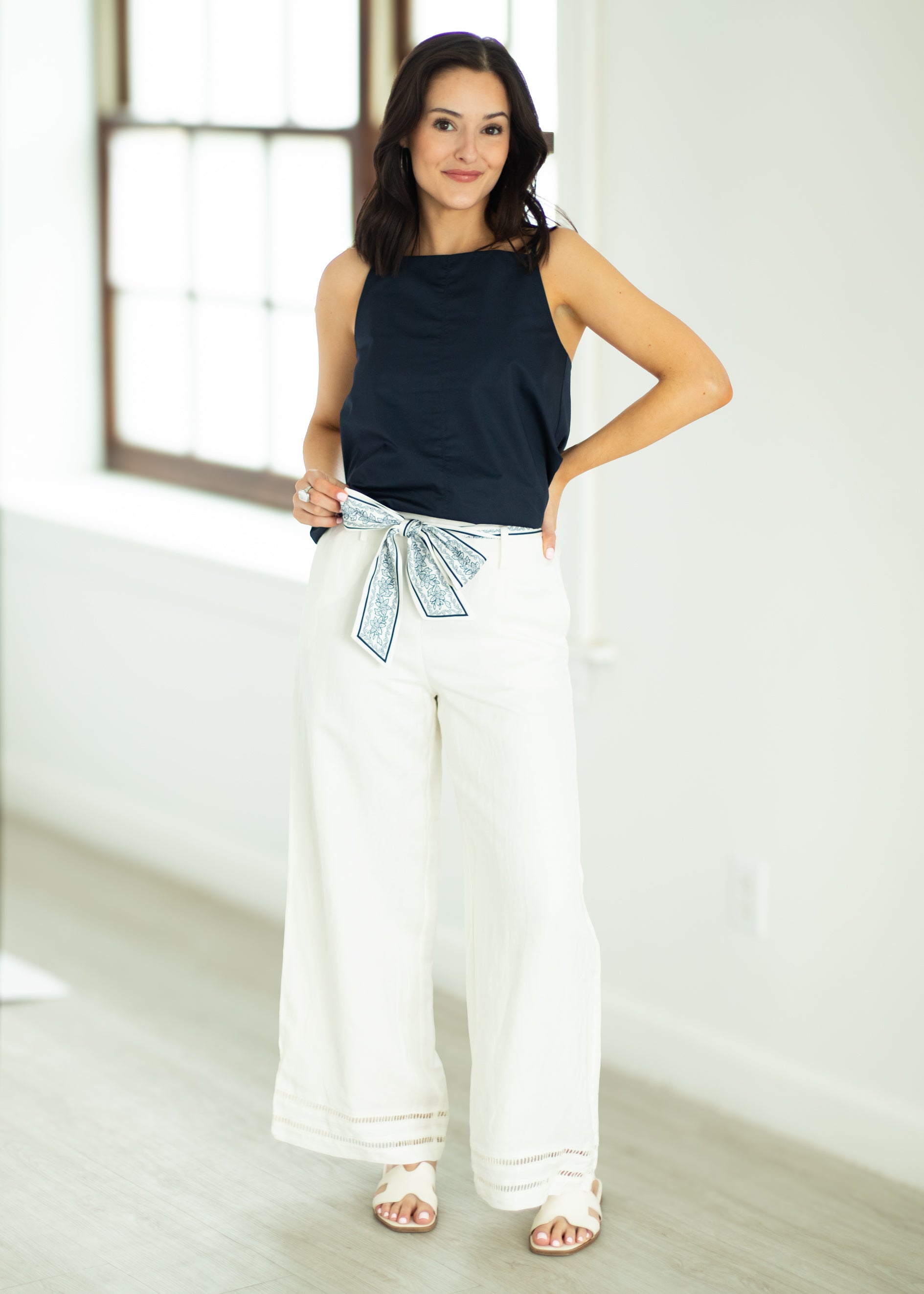 Woman wearing a black sleeveless top and white pants with a decorative belt in a bright room.