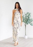 Woman wearing a floral maxi dress in a minimalistic setting with a plant in the background.