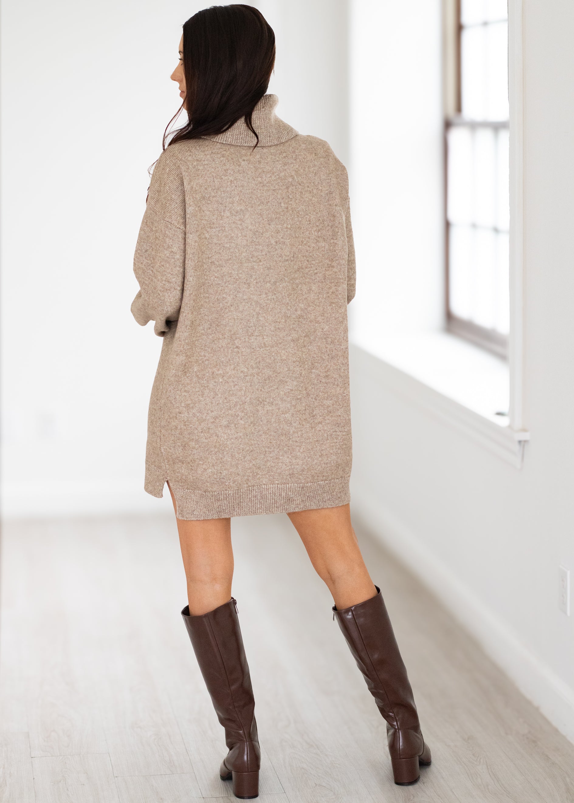 Woman wearing a beige sweater dress and brown knee-high boots in a minimalistic room.