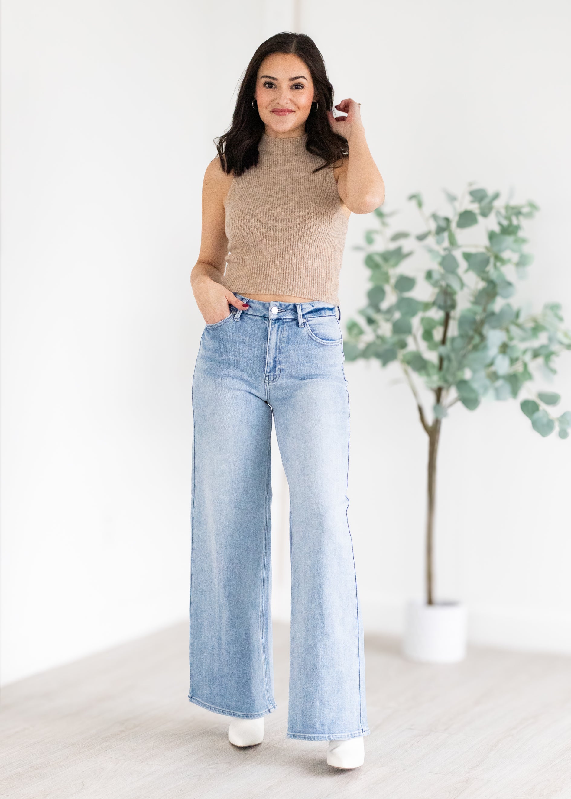 Woman wearing a beige sleeveless top and light blue jeans standing indoors with a plant in the background.