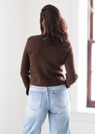 Person wearing a brown sweater and light blue jeans standing against a white wall.