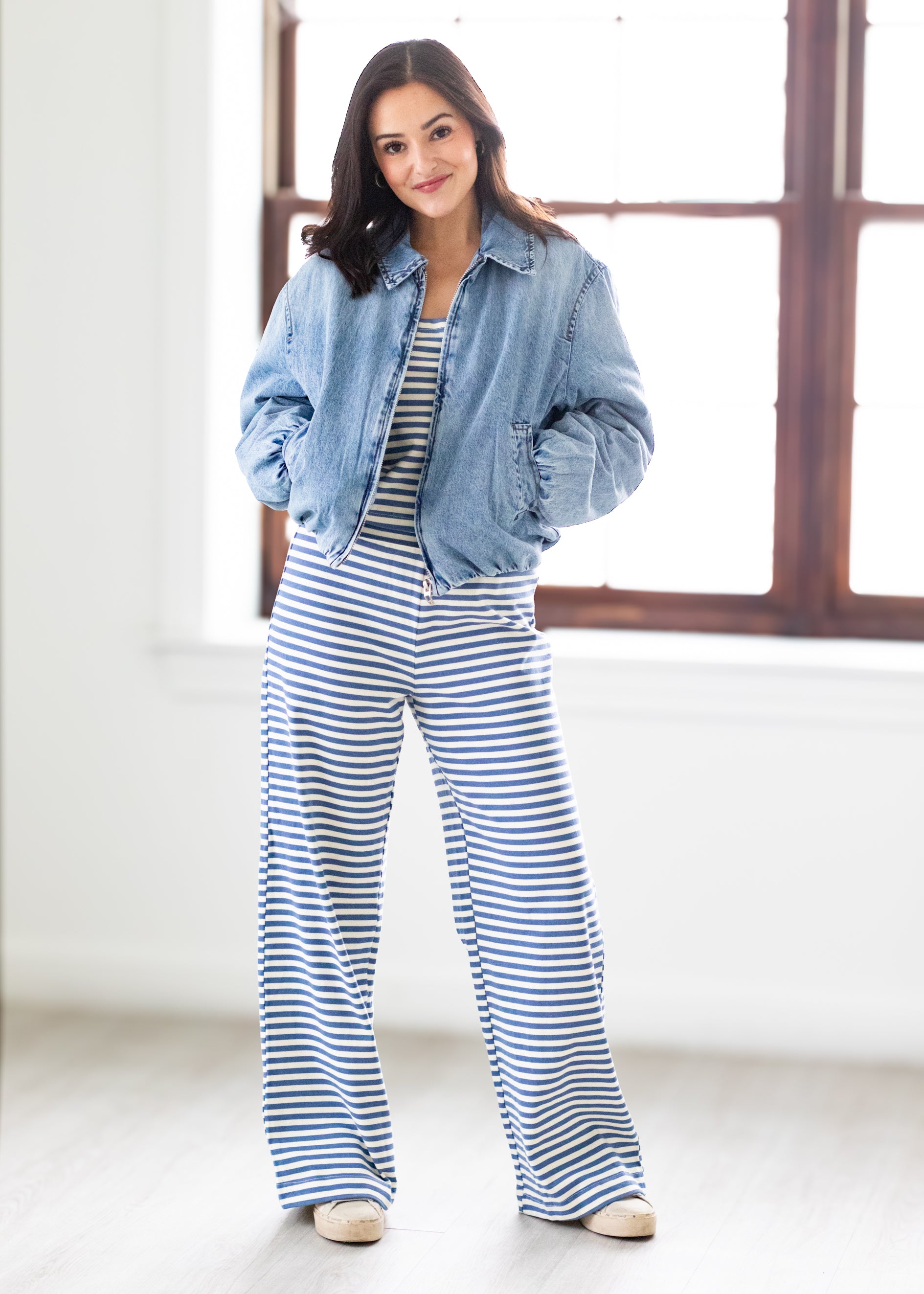 Woman wearing a denim jacket and blue and white striped pants standing in a bright room.