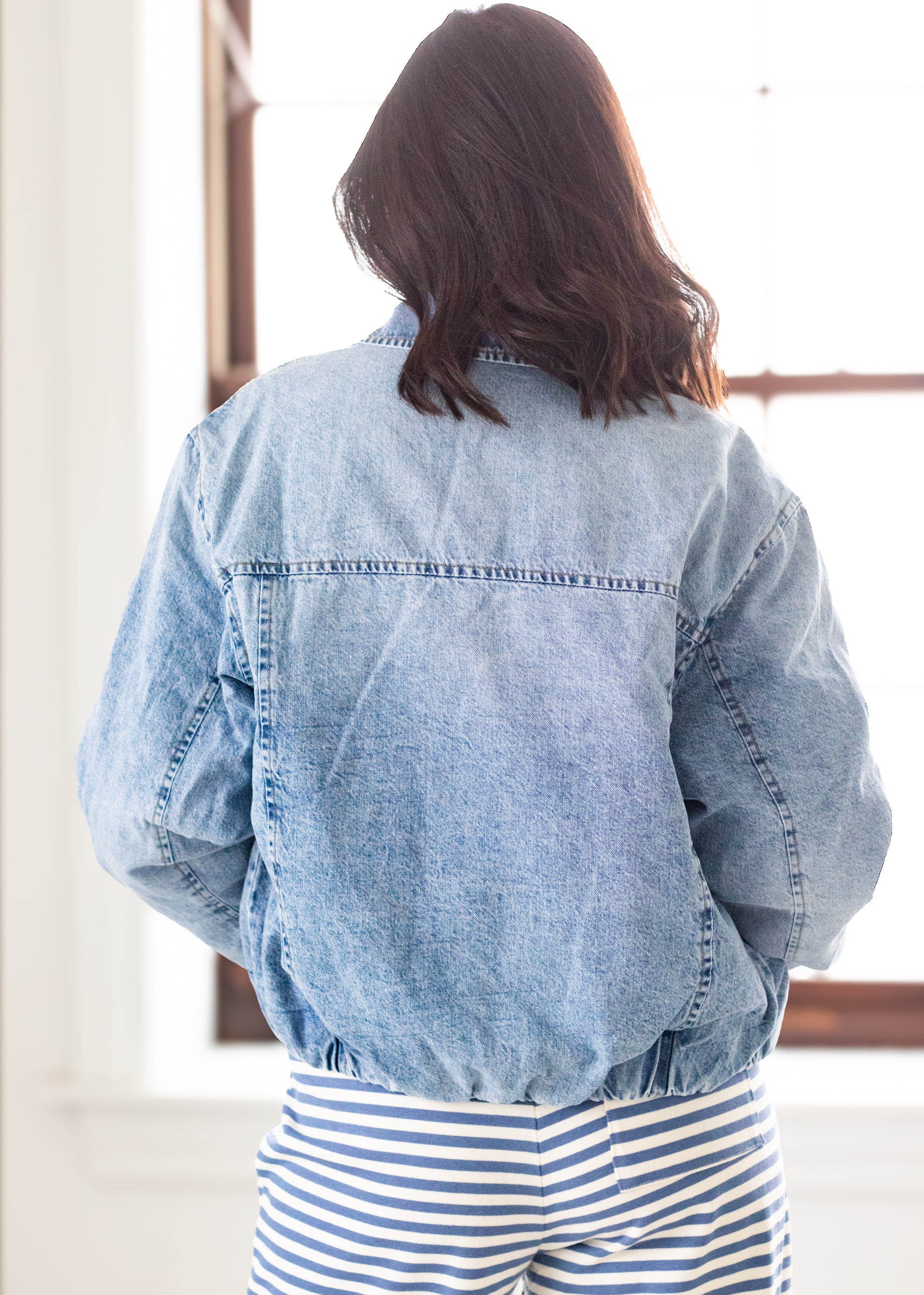 Person wearing a denim jacket and striped pants in a room with a window.