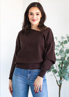 Woman wearing a brown sweater and blue jeans standing against a white background with a plant.