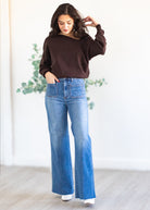 Woman wearing a brown sweater and blue jeans standing indoors with a light background