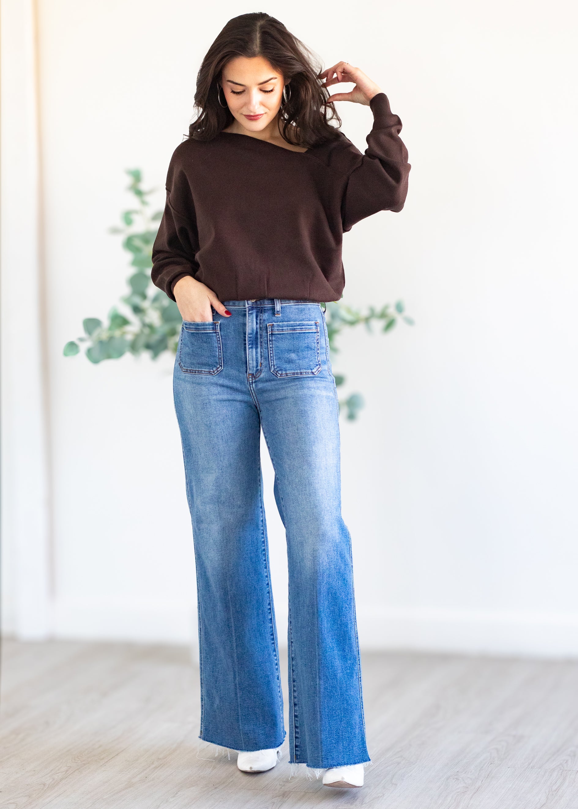 Woman wearing a brown sweater and blue jeans standing indoors with a light background