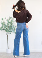 Person wearing a brown sweatshirt and blue jeans standing in a minimalistic room with a plant.