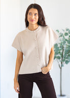 Woman wearing a beige blouse with short sleeves and buttons, standing against a white background.