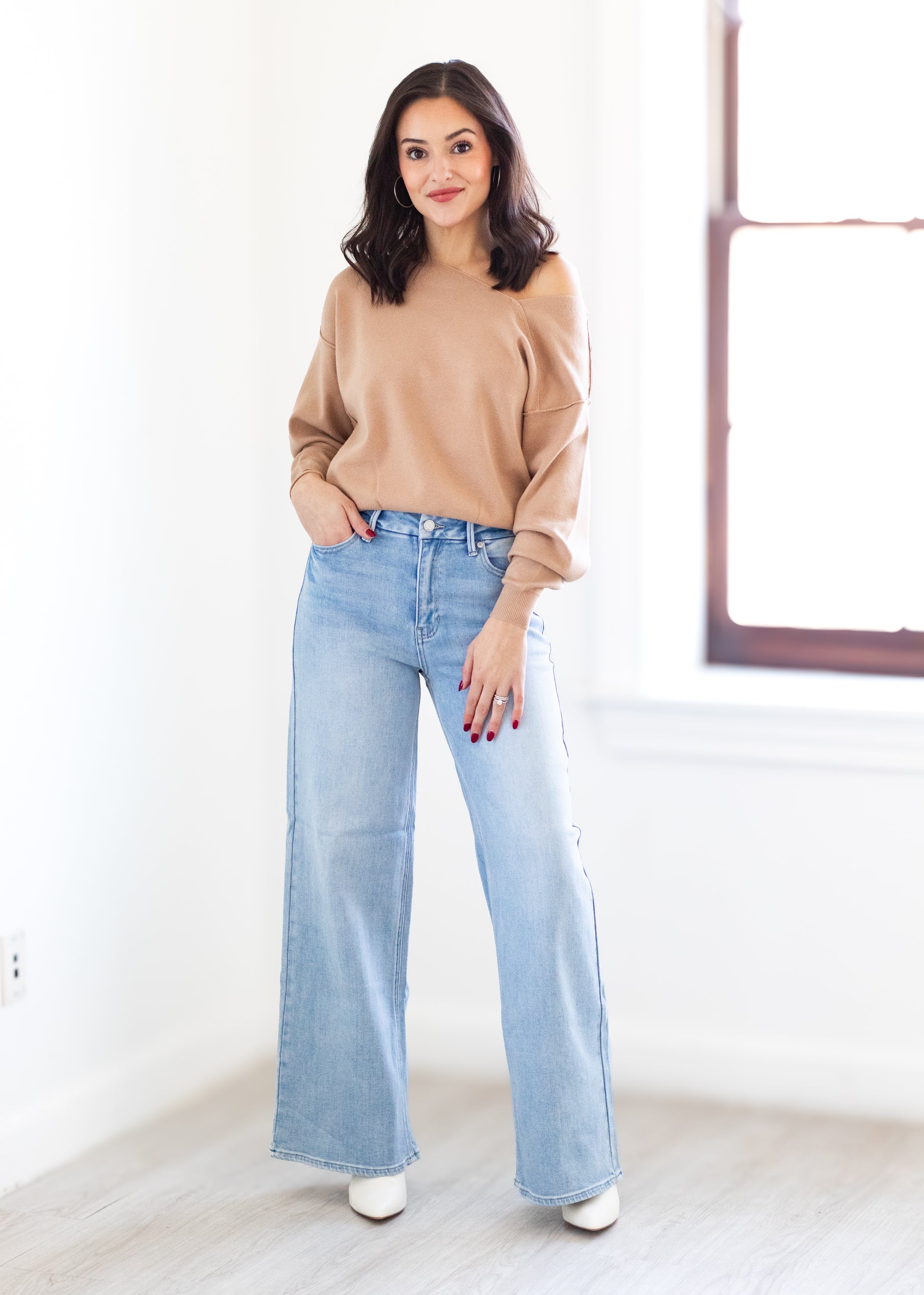 Woman wearing a beige sweater and light blue jeans standing in a room with a white wall and wooden floor.
