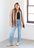 Woman wearing a beige faux fur coat, black top, and light blue jeans indoors.
