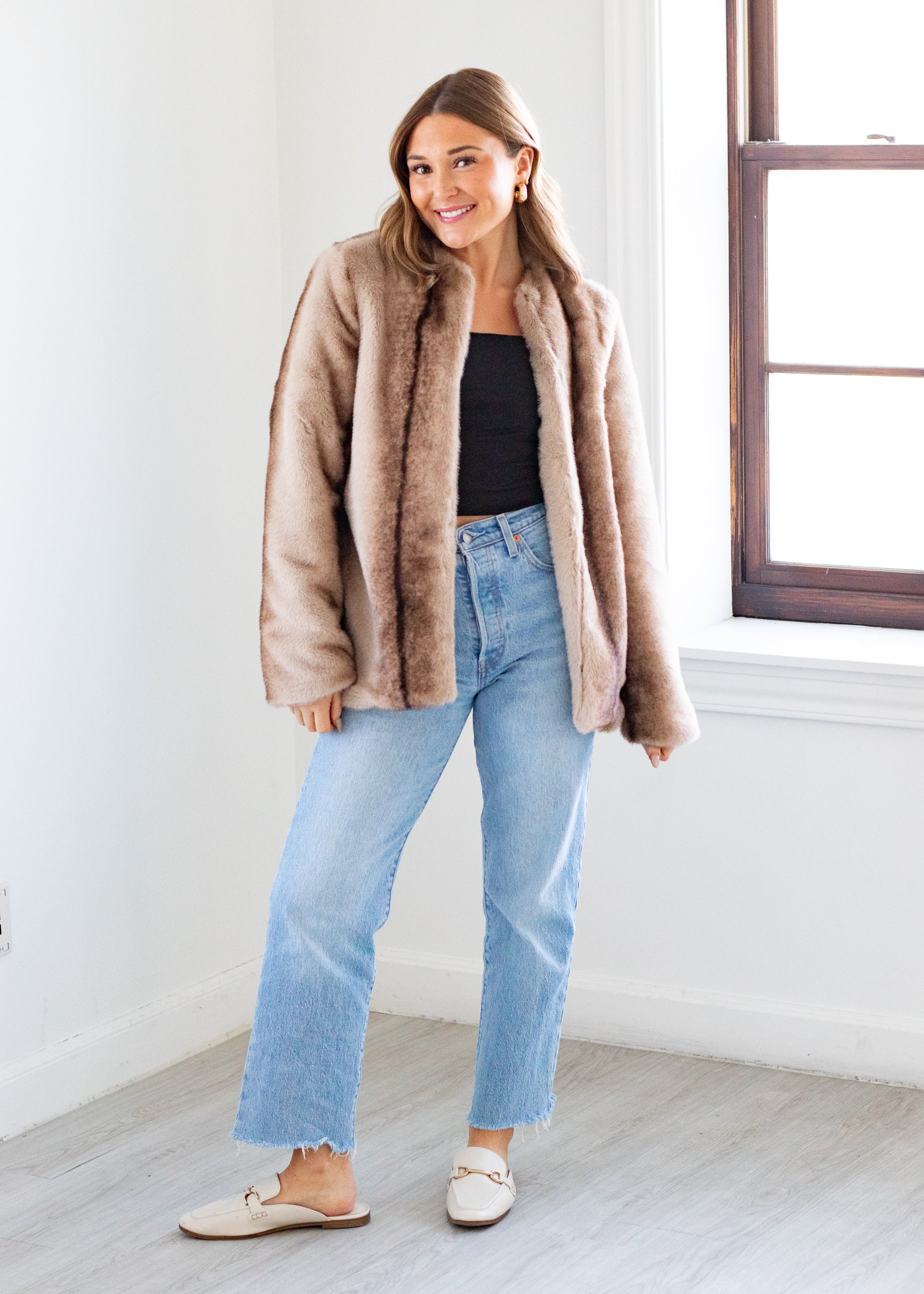 Woman wearing a beige faux fur coat, black top, and light blue jeans indoors.