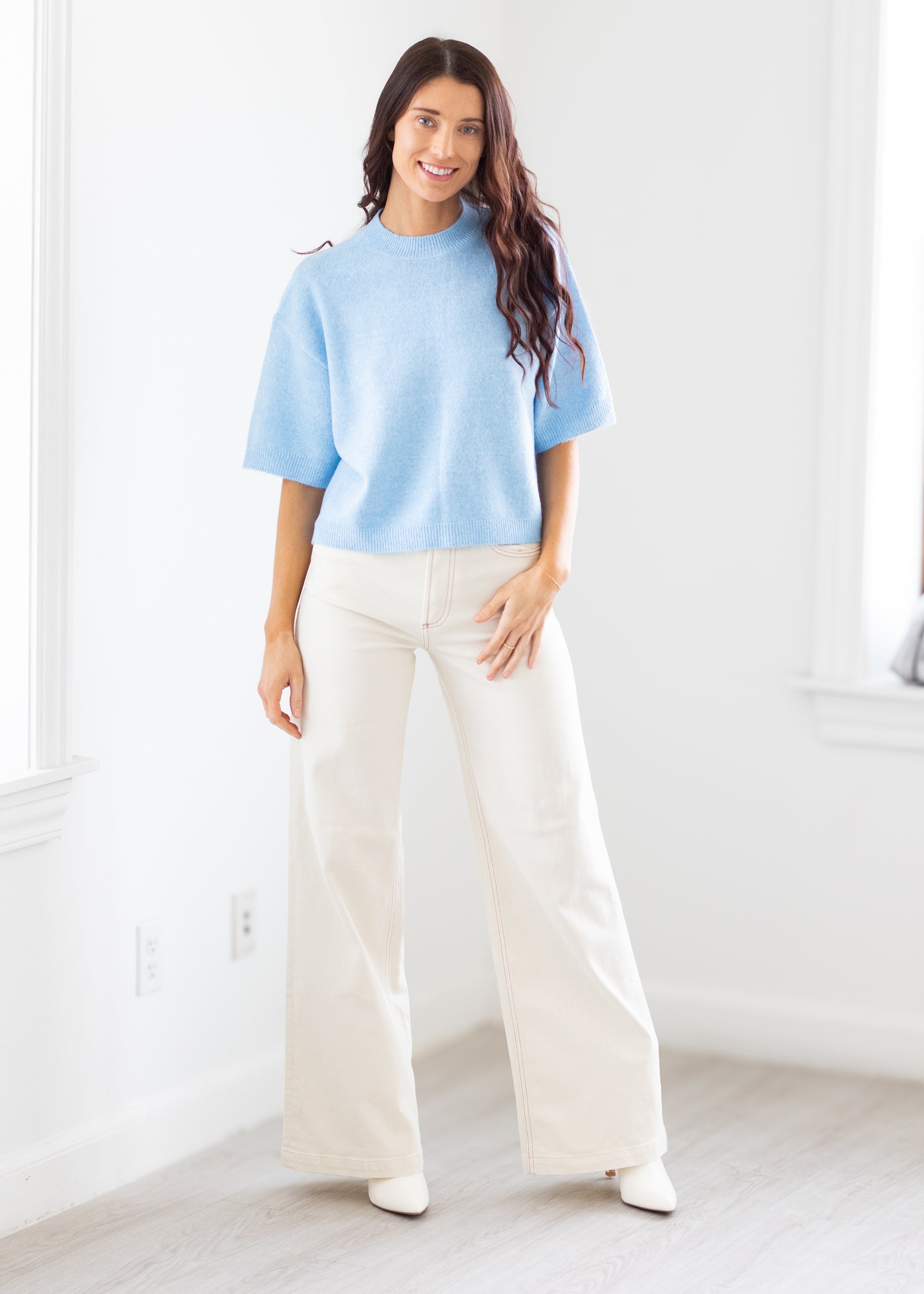 Woman wearing a light blue sweater and white pants in a bright room.