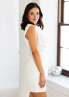 Woman wearing a white dress standing against a light-colored wall.