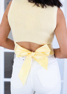 Person wearing a yellow sleeveless top with a bow detail and white pants.