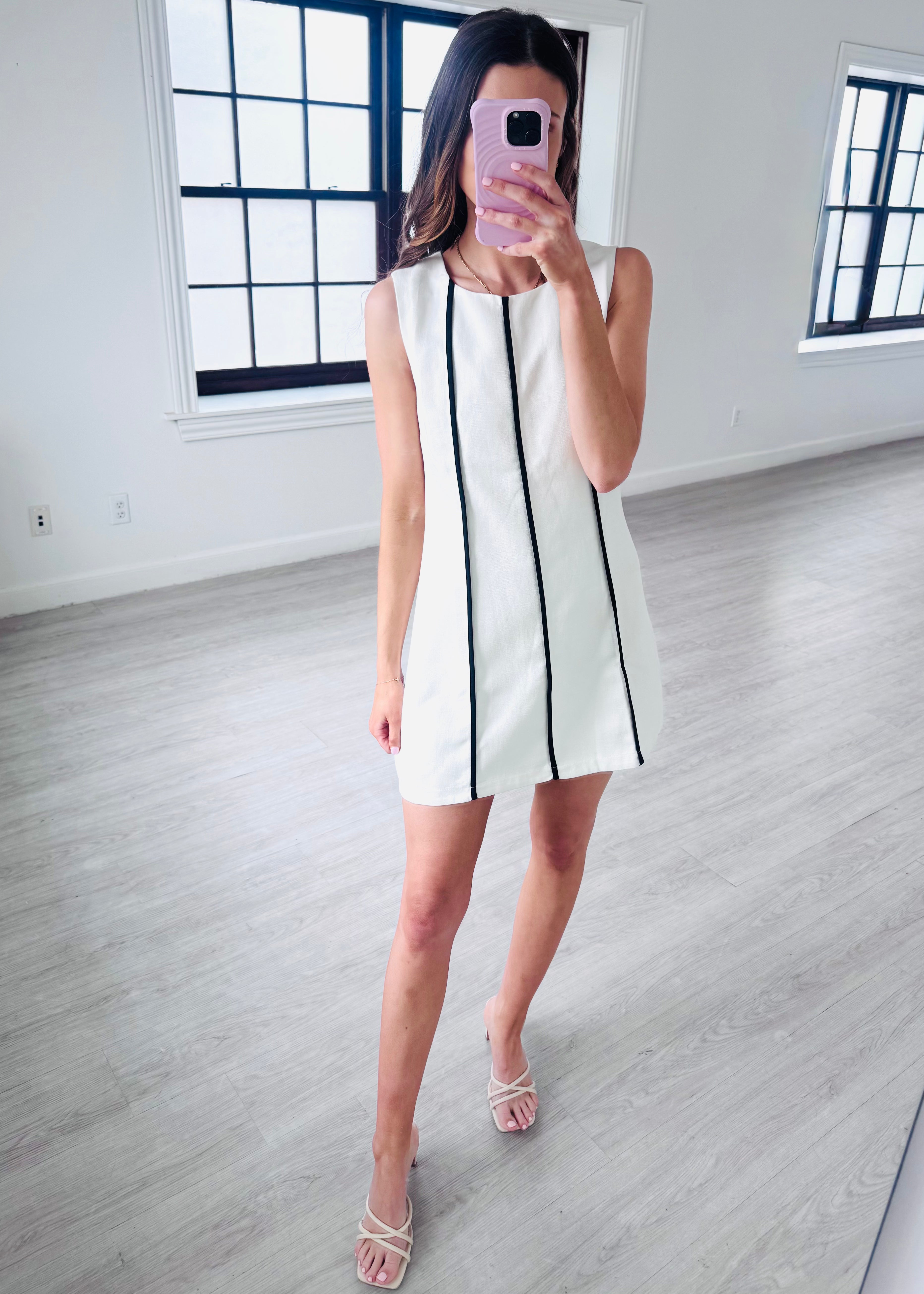 Woman taking a mirror selfie wearing a white dress with black stripes in a minimalistic room.