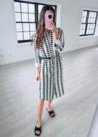 Woman taking a mirror selfie wearing a black and white patterned dress in a minimalistic room.