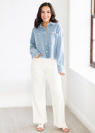 Woman wearing a light blue denim jacket and white pants standing in a room with a neutral background.