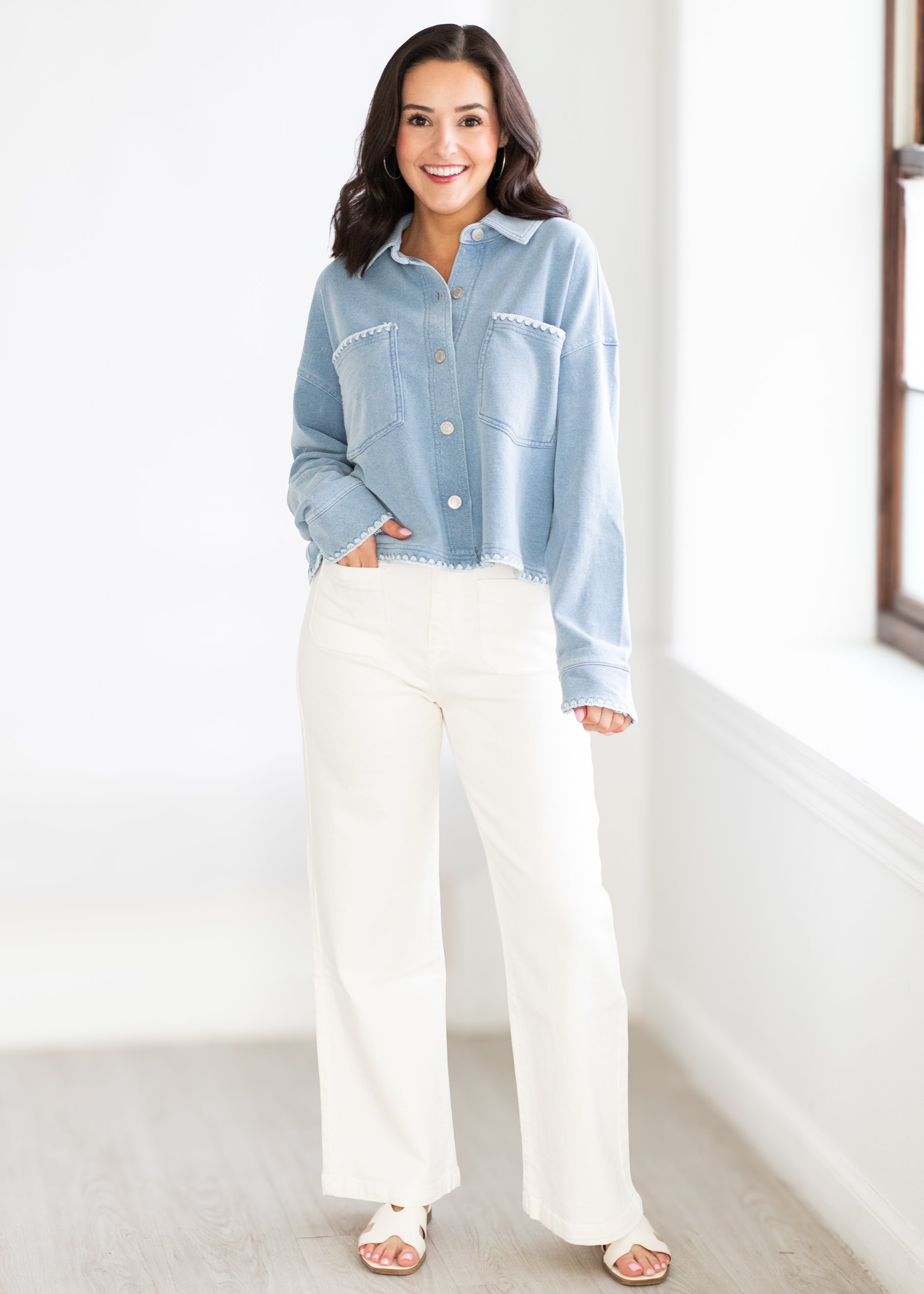 Woman wearing a light blue denim jacket and white pants standing in a room with a neutral background.