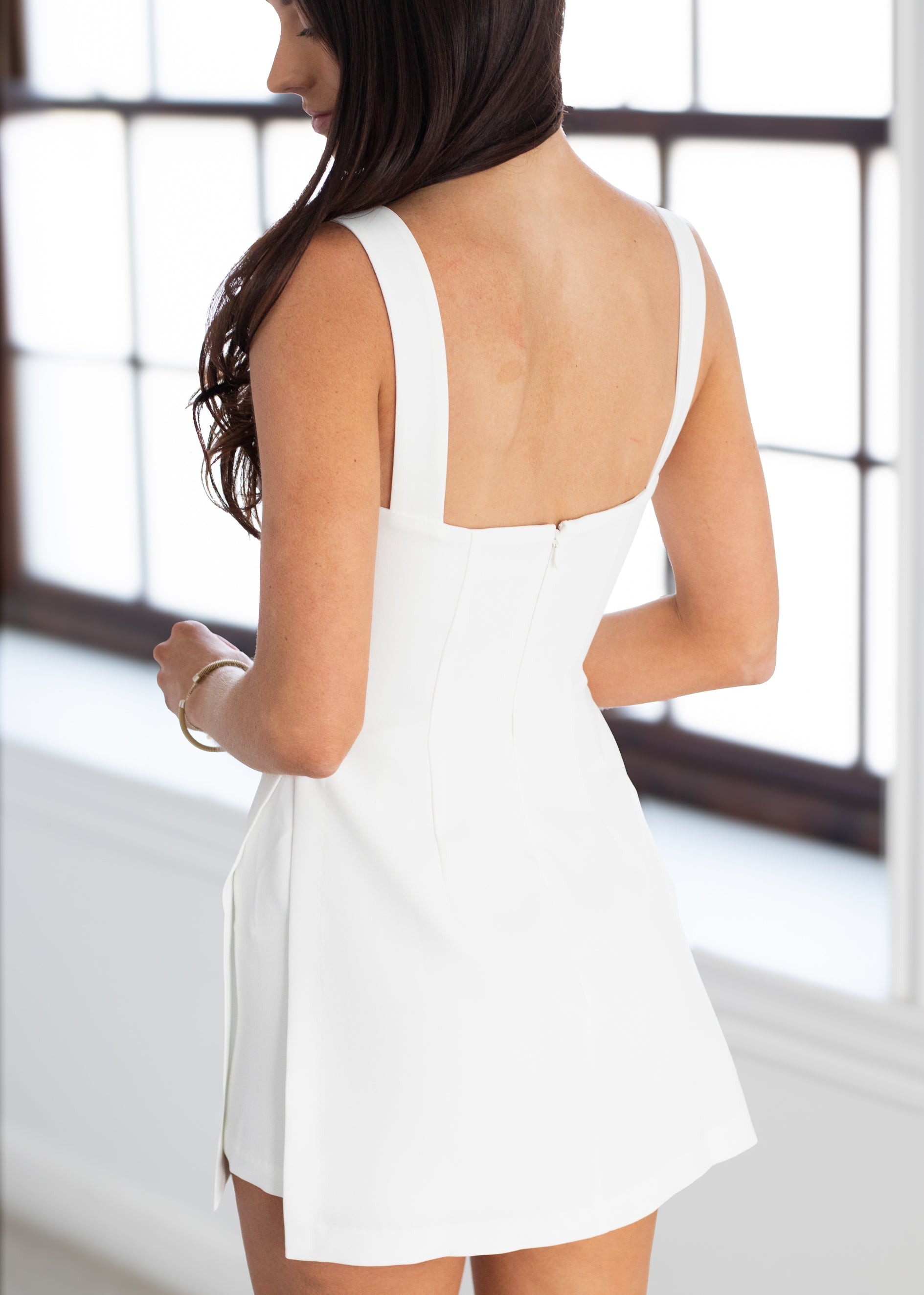 Woman wearing a white dress with a backless design, standing in front of a window.