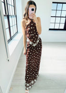 A woman modeling a halter-neck maxi dress in a dark brown color with a white bohemian-style print and patterned hem, set against a plain light gray background.