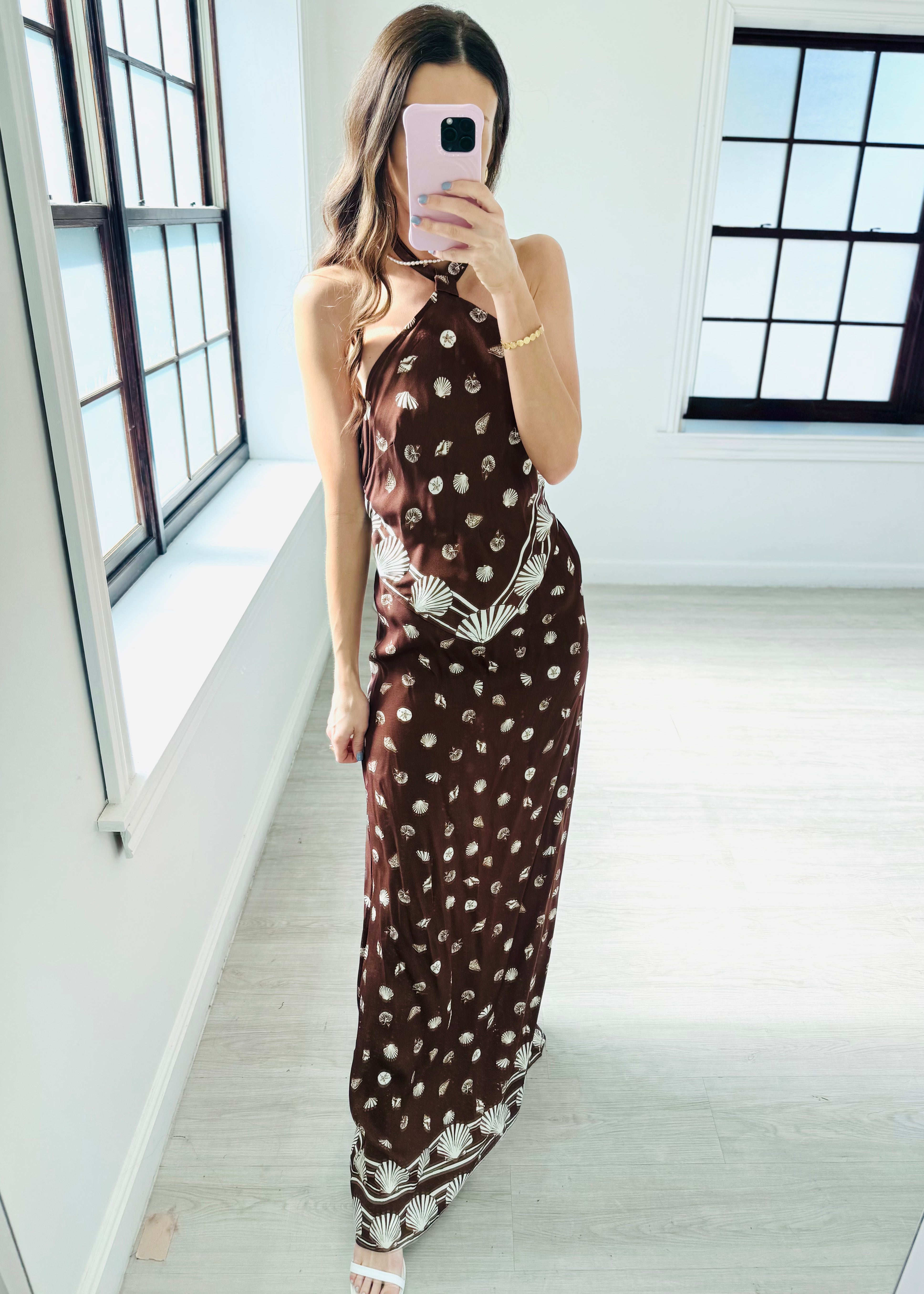 A woman modeling a halter-neck maxi dress in a dark brown color with a white bohemian-style print and patterned hem, set against a plain light gray background.