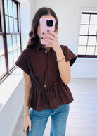 A woman takes a mirror selfie wearing a dark brown short-sleeve button-up blouse with a drawstring waist and blue jeans in a bright room with large windows.