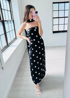 A woman takes a mirror selfie in a bright room wearing a black strapless maxi dress with white polka dots and a matching black choker.