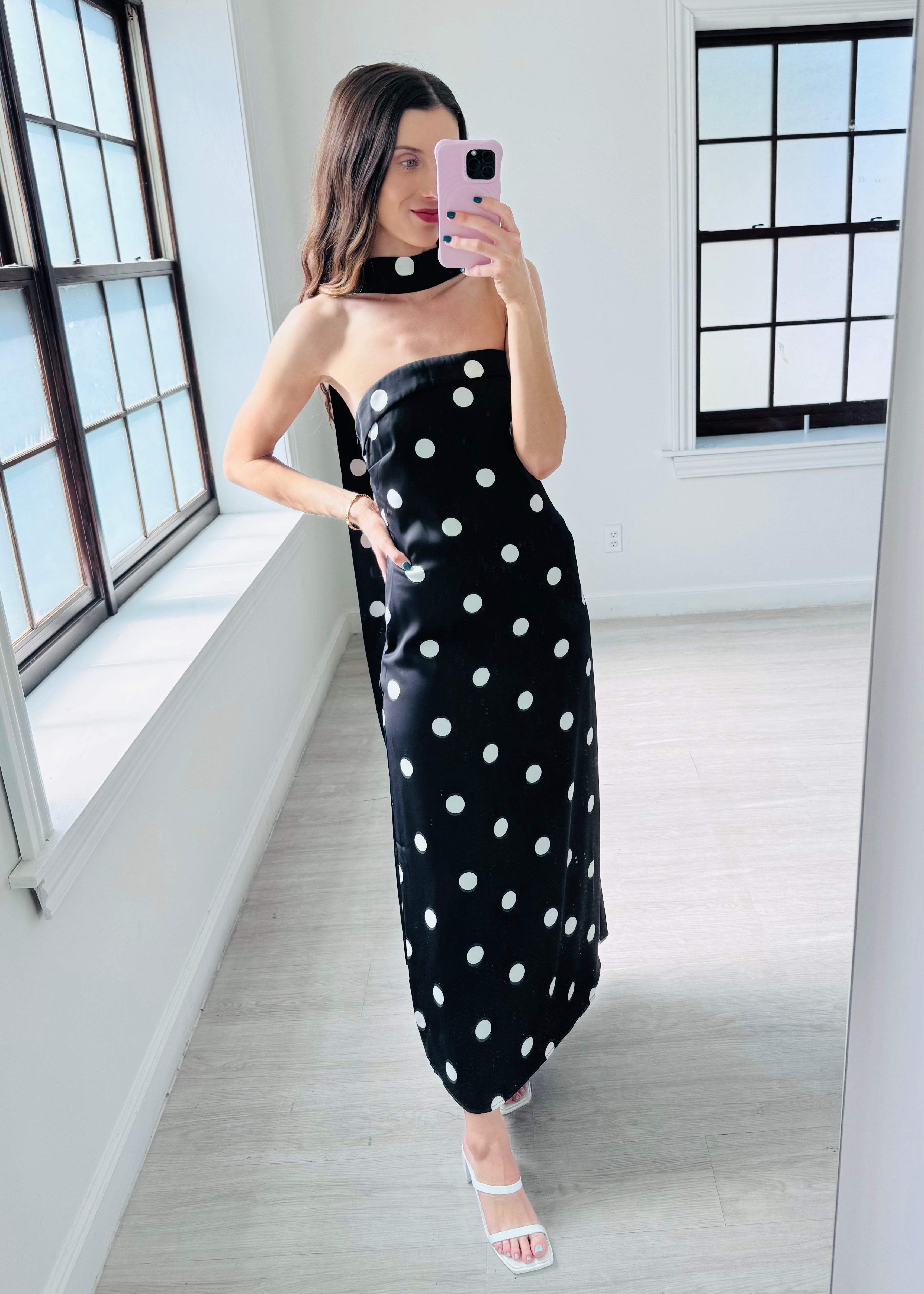 A woman takes a mirror selfie in a bright room wearing a black strapless maxi dress with white polka dots and a matching black choker.