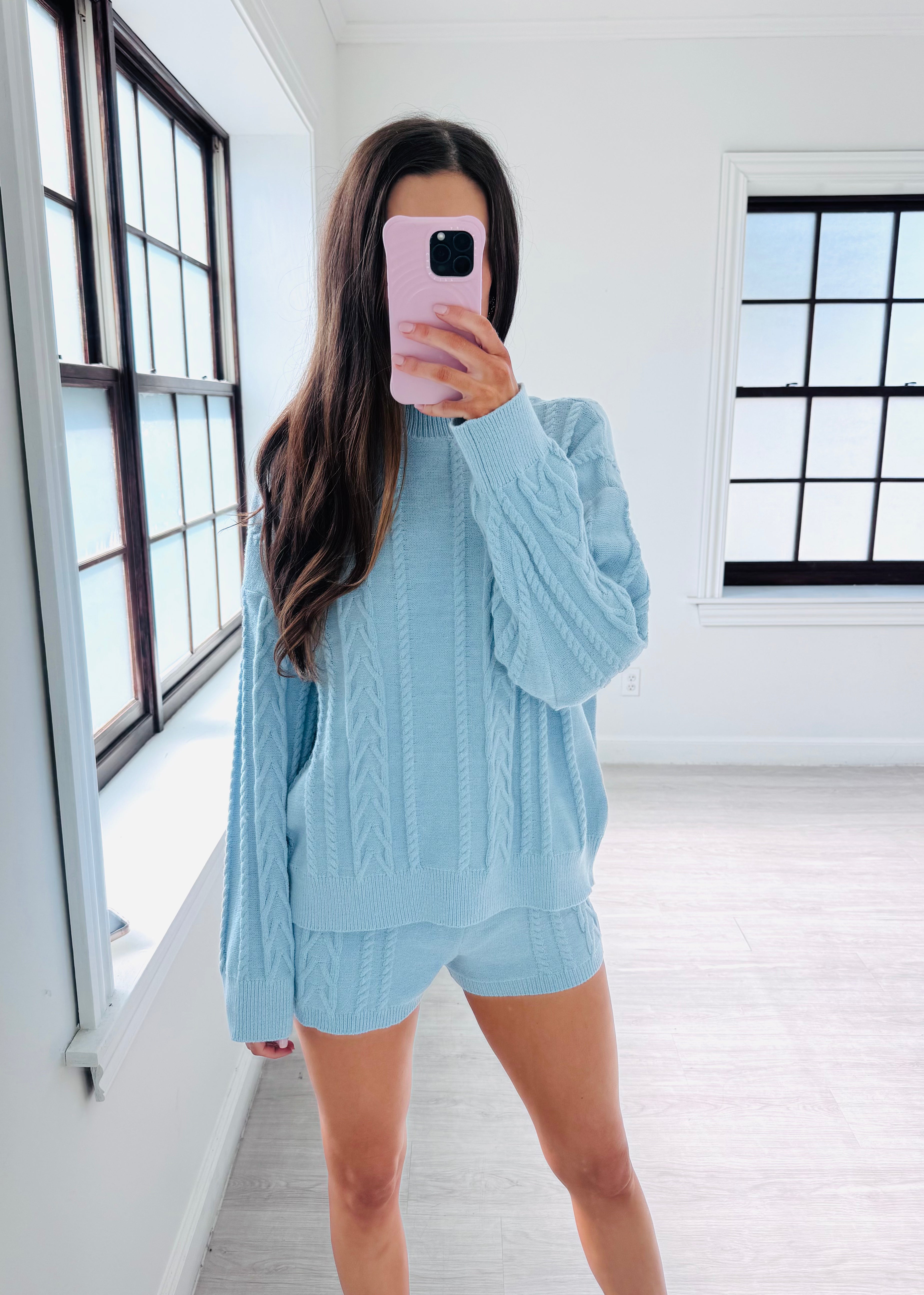 A woman takes a mirror selfie in a bright room wearing a light blue cable-knit matching lounge set consisting of a long-sleeve top and shorts.