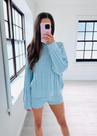 A woman takes a mirror selfie in a bright room wearing a light blue cable-knit matching lounge set consisting of a long-sleeve top and shorts.