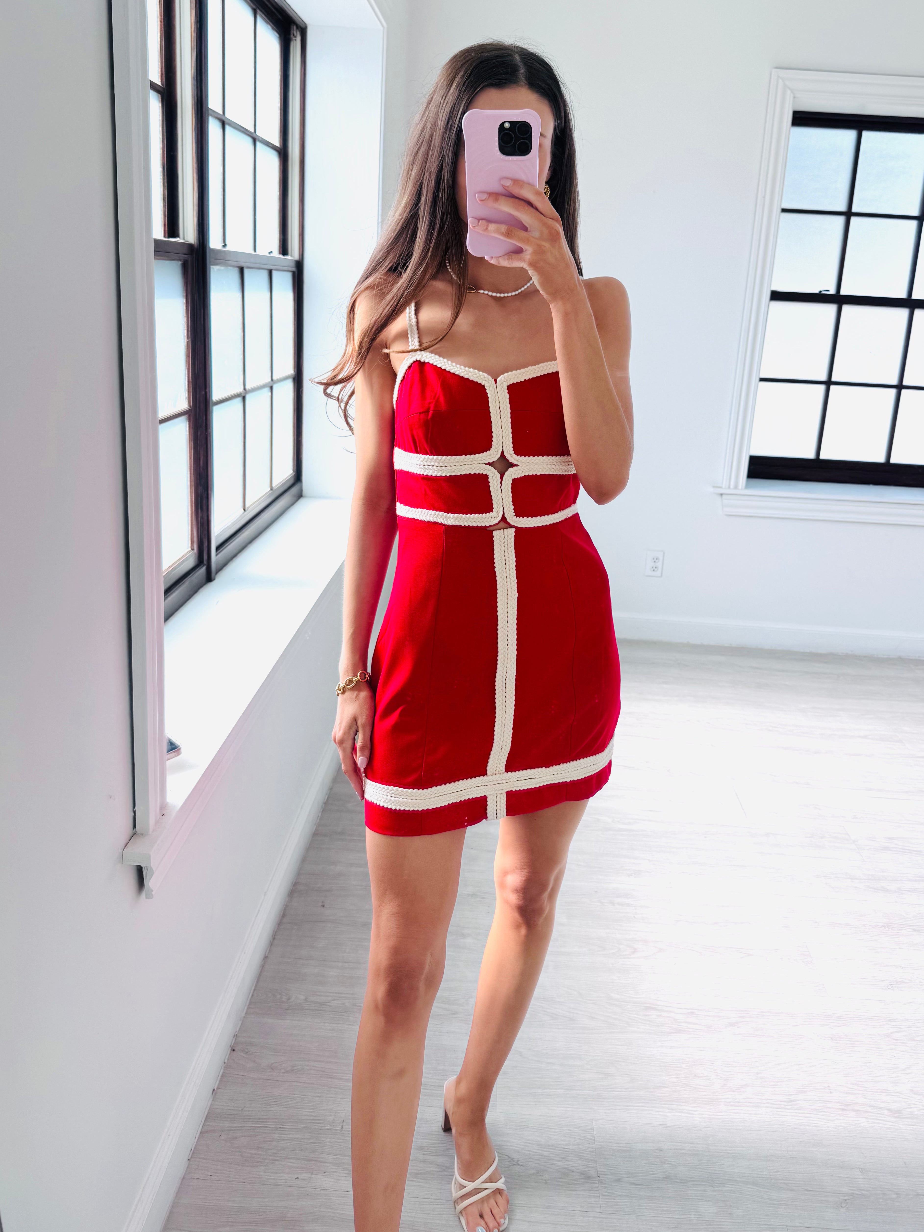 A woman takes a mirror selfie in a bright room wearing a red mini dress with white contrast trim and straps. The dress features a structured, geometric design with a central vertical panel and horizontal bands.