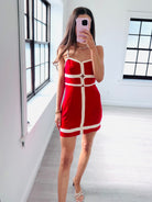 A woman takes a mirror selfie in a bright room wearing a red mini dress with white contrast trim and straps. The dress features a structured, geometric design with a central vertical panel and horizontal bands.