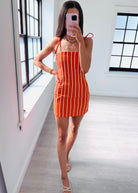 A woman takes a mirror selfie in a bright room wearing a bright orange and white vertically striped mini dress with thin shoulder straps.