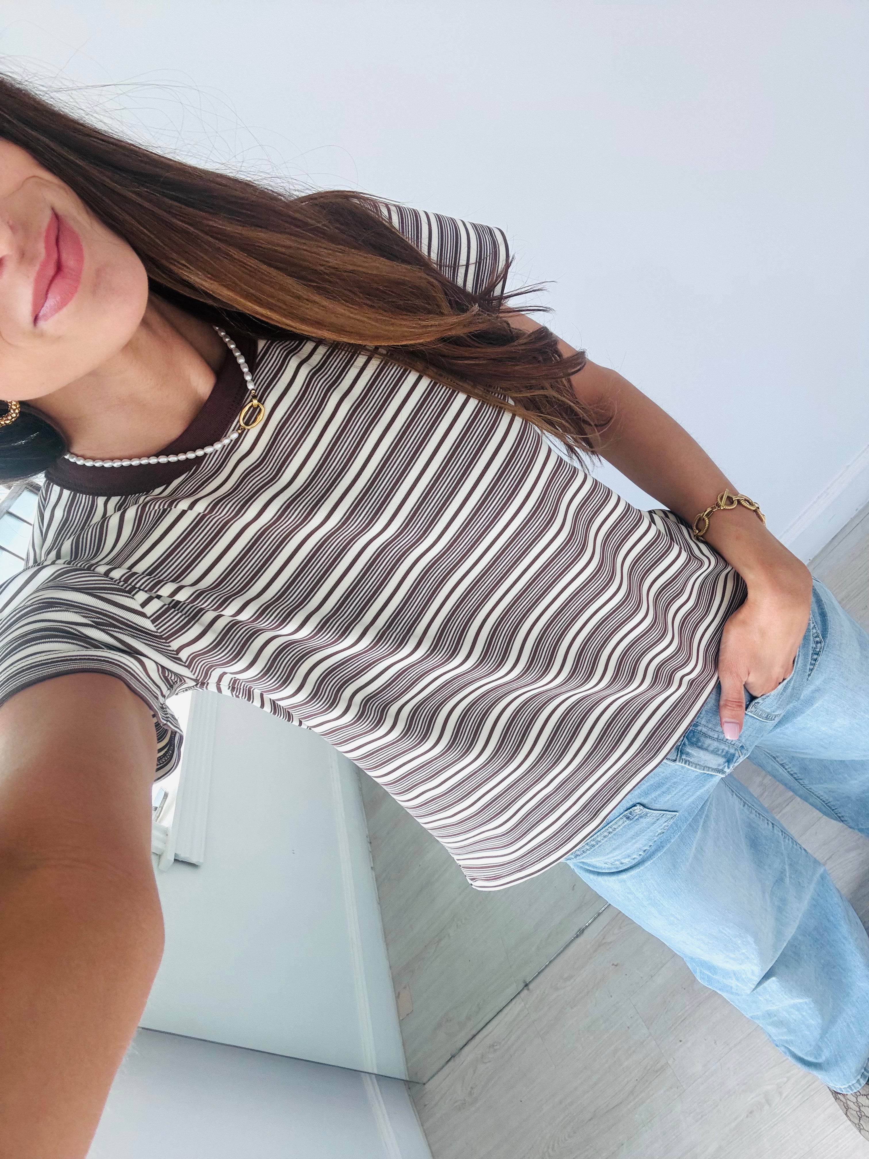A woman wearing a brown, white, and gray striped short-sleeve t-shirt with a dark brown collar, paired with light blue jeans.
