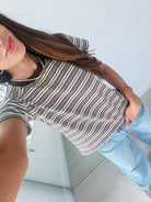 A woman wearing a brown, white, and gray striped short-sleeve t-shirt with a dark brown collar, paired with light blue jeans.