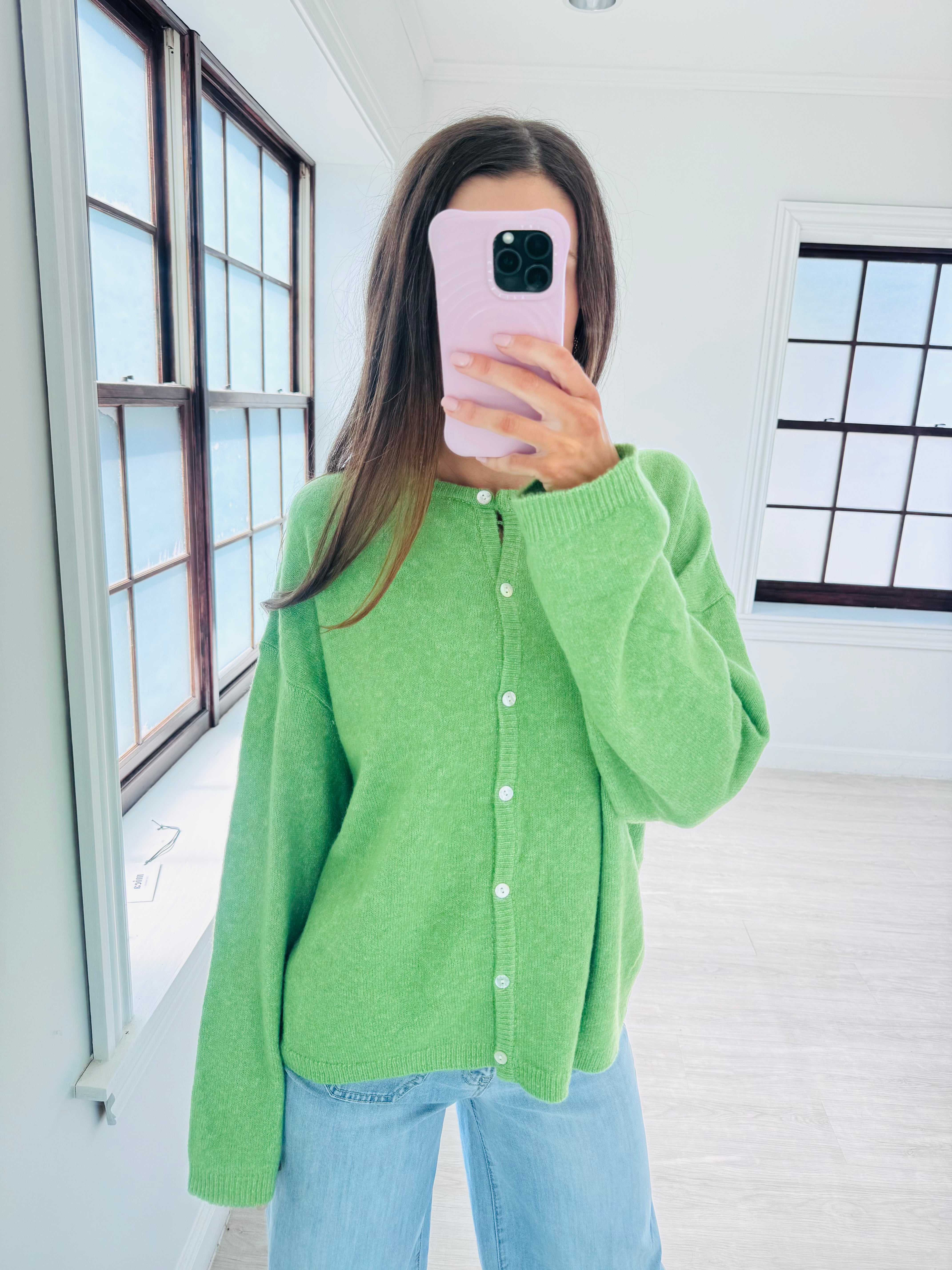 A woman takes a mirror selfie wearing a bright lime green knit cardigan with white buttons and light blue jeans in a bright, modern room with large windows.