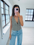 A woman takes a mirror selfie wearing a black and white striped halter top and light blue jeans in a bright, modern room with large black-framed windows.
