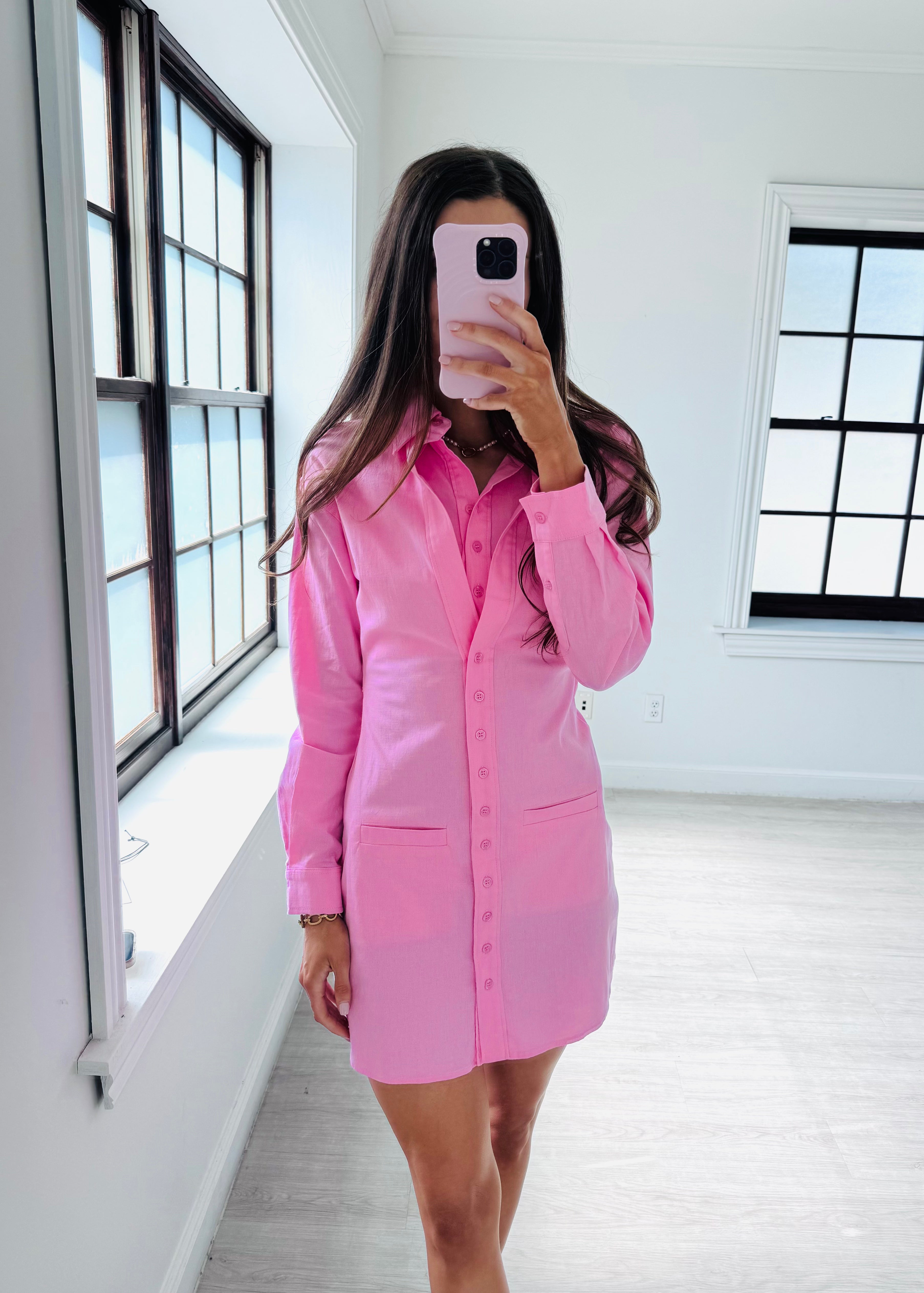 A woman takes a mirror selfie in a bright, modern room wearing a pink button-down shirt dress. The dress has long sleeves, a pointed collar, and front patch pockets.
