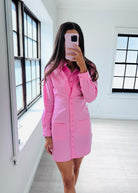 A woman takes a mirror selfie in a bright, modern room wearing a pink button-down shirt dress. The dress has long sleeves, a pointed collar, and front patch pockets.