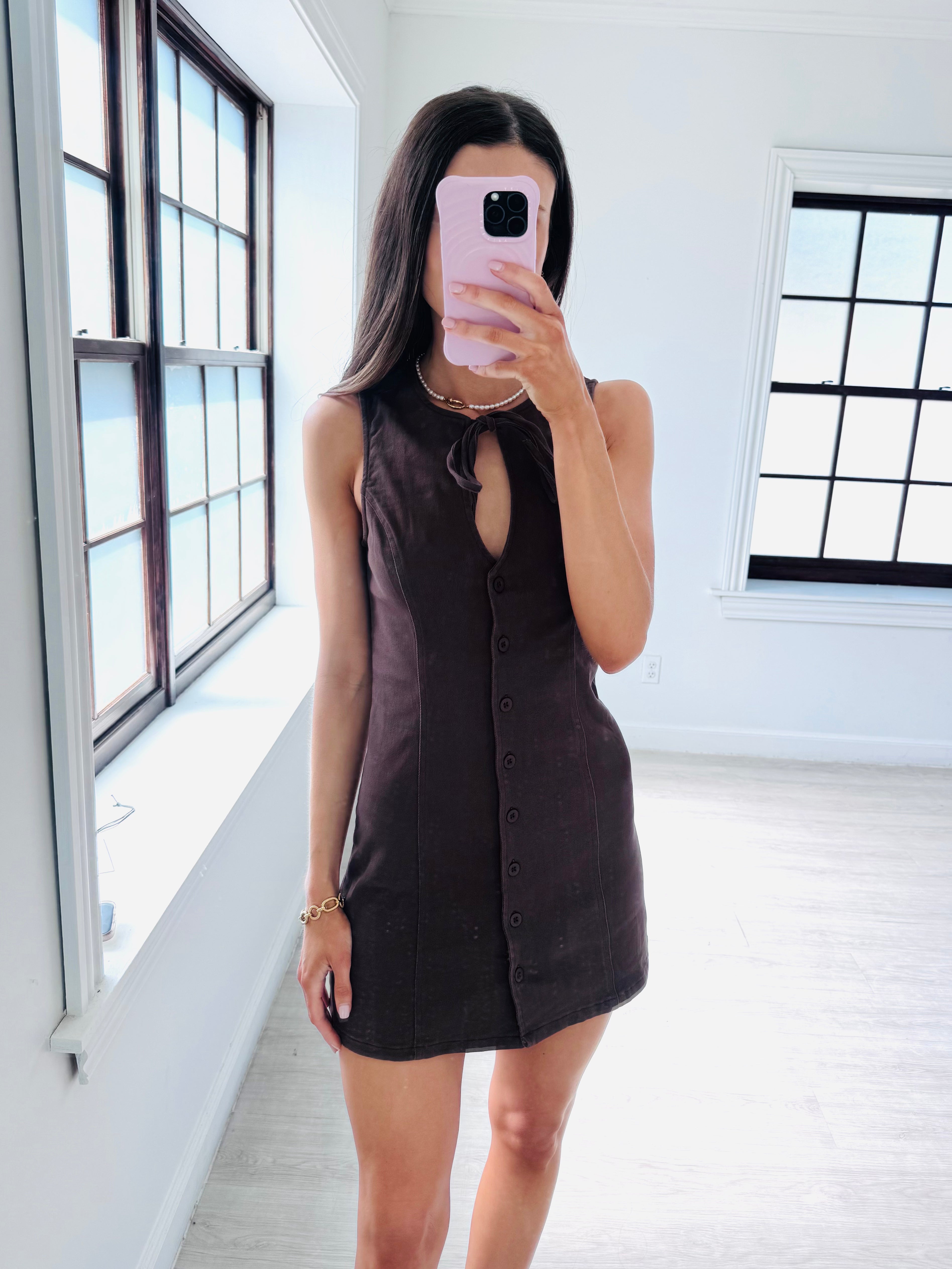 A woman takes a mirror selfie in a bright room wearing a sleeveless dark brown button-front mini dress with a tie-neck detail. She is holding a light pink smartphone.